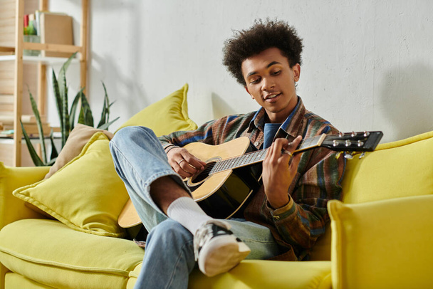 A young African American man playing an acoustic guitar on a yellow couch while talking on his phone. - Fotoğraf, Görsel