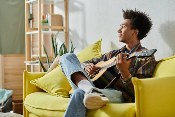A young African American man plays an acoustic guitar while seated on a yellow couch. - 写真・画像