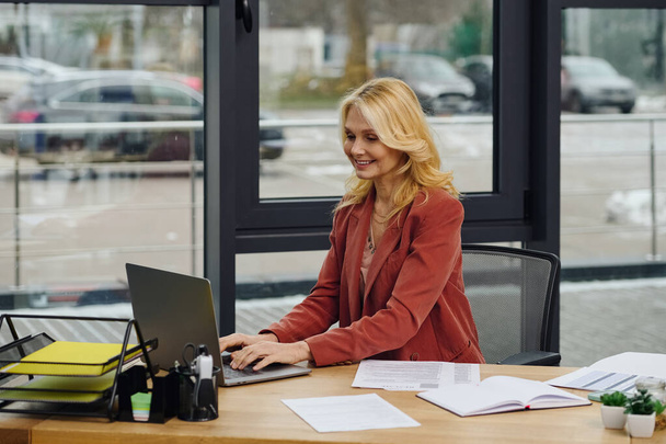Μια γυναίκα κάθεται σε ένα γραφείο με ένα φορητό υπολογιστή, επικεντρωμένη στη δουλειά της.. - Φωτογραφία, εικόνα