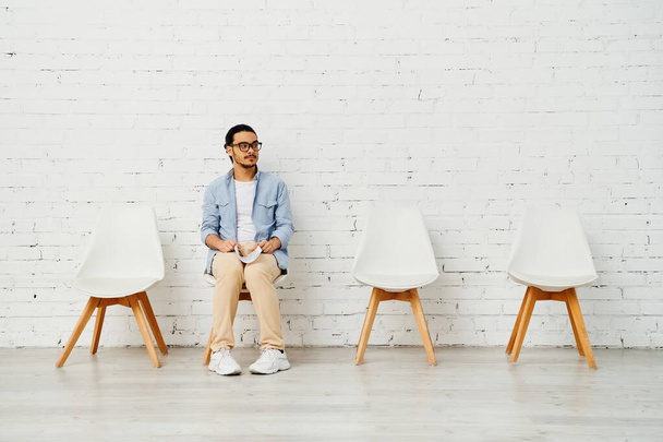 Ένας άντρας κάθεται σε μια σειρά από καρέκλες σε έναν λευκό τοίχο από τούβλα.. - Φωτογραφία, εικόνα