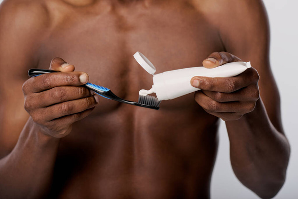 Um afro-americano segurando uma escova de dentes e pasta de dentes em um estúdio com um fundo branco. - Foto, Imagem