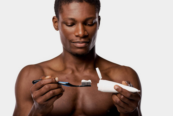 Hemdloser afroamerikanischer Mann im Studio mit weißem Hintergrund, der eine Zahnbürste und eine Zahnbürste in der Hand hält. - Foto, Bild