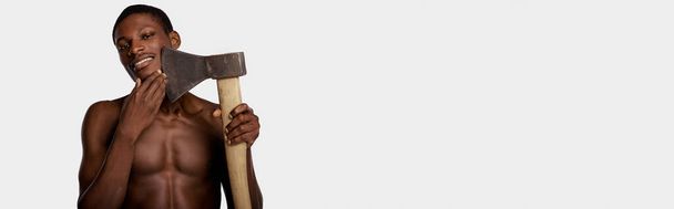 Un afroamericano sin camisa sostiene un gran hacha en su mano derecha contra un fondo de estudio blanco. - Foto, Imagen