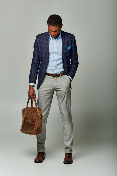 Afroamerikaner im karierten Blazer hält eine braune Tasche vor einem glatten grauen Hintergrund. - Foto, Bild
