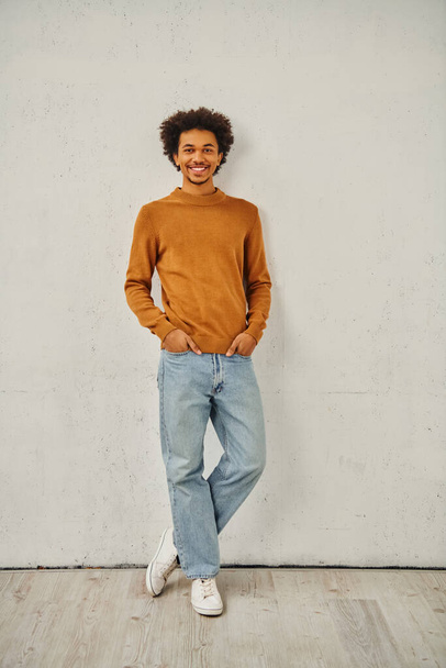 Młody człowiek w brązowym swetrze i dżinsach stojący z przemyślanym wyrazem twarzy pod ścianą. - Zdjęcie, obraz