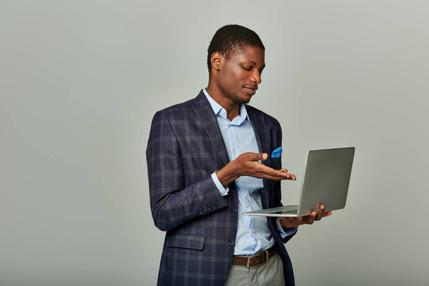 Jeune homme d'affaires afro-américain dans un blazer à carreaux fonctionnant ordinateur portable sur fond gris. - Photo, image