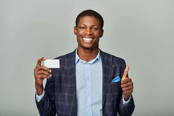 Joven hombre de negocios afroamericano en chaqueta a cuadros sosteniendo una tarjeta de visita y mostrando un gesto de pulgares hacia arriba. - Foto, imagen