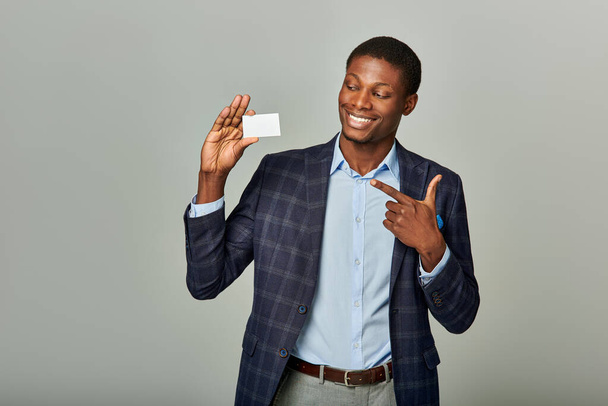 Hezký africký podnikatel ve stylovém obleku elegantně drží vizitku na šedém pozadí. - Fotografie, Obrázek