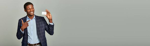 Africký americký podnikatel v kostkovaném blejzru drží vizitku na šedém pozadí. - Fotografie, Obrázek