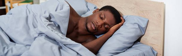 Młody Afroamerykanin spokojnie śpiący w łóżku pod przytulnym niebieskim kocem, gotowy obudzić się rano. - Zdjęcie, obraz
