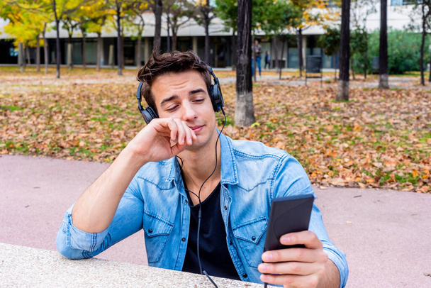 Mladý muž v džínové bundě používá sluchátka a smartphone venku, opírající se o lavičku v parku, uvolněný a soustředěný. - Fotografie, Obrázek