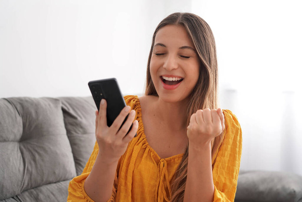 Jeune femme excitée avec les yeux fermés et le poing levé tient smartphone sur le canapé à la maison - Photo, image