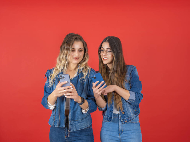 Due donne che usano il telefono, ritratto di giovani ventenni due donne che usano il telefono. Sfondo rosso. Puntando dito touch screen, sorridente mostrando buone notizie, social media, raccomandando nuovo concetto di applicazione cool. - Foto, immagini