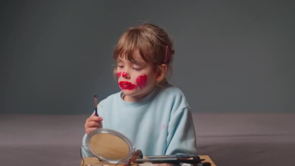 Una graziosa bambina seduta davanti a uno specchio cerca di truccarsi immergendo un pennello nei cosmetici. - Filmati, video