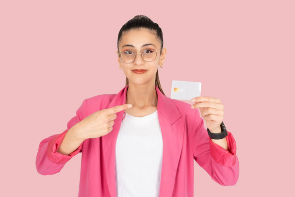 Pointing credit card, portrait of young caucasian brunette woman wear glasses and pink formal jacket holding pointing credit card. Recommending online shopping bank services standing pink background. - Photo, Image