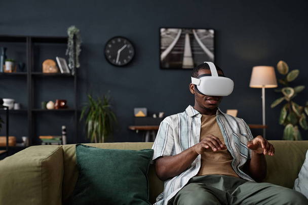 Młody czarny mężczyzna noszący słuchawki VR siedzący na kanapie w salonie wpisujący na wirtualnej klawiaturze, przestrzeń do kopiowania - Zdjęcie, obraz
