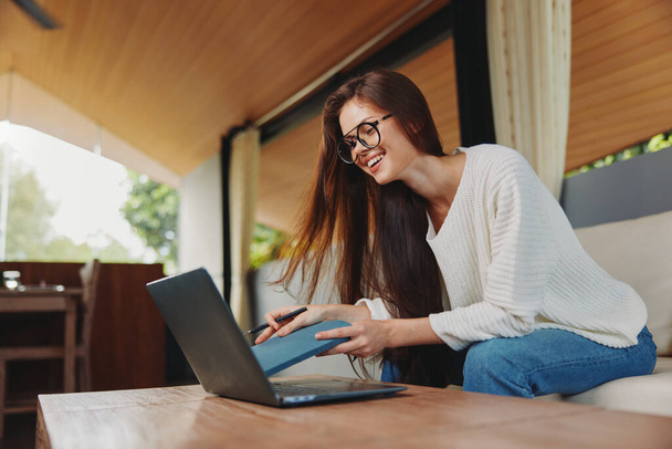 Uśmiechnięta kobieta pracująca na laptopie w wygodnym biurze domowym na sofie Jest nauczycielką prowadzącą zajęcia online, noszącą okulary i używającą notebooka do edukacji Salon zapewnia - Zdjęcie, obraz