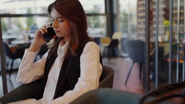 Een jonge professionele vrouw houdt zich bezig met een serieus telefoongesprek terwijl ze in een stijlvolle, eigentijdse café-omgeving zit. Professionele vrouw het maken van een zakelijke oproep in een modern café - Video
