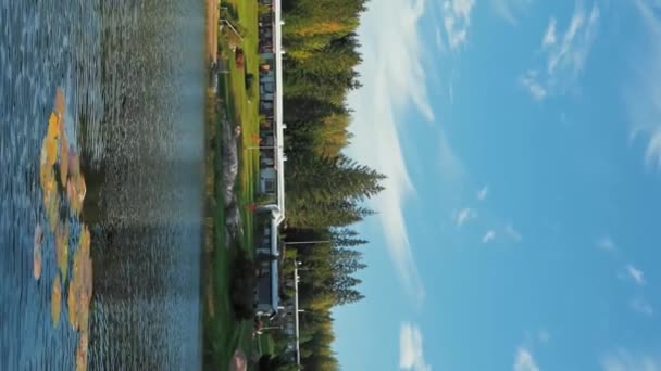 Εξοχικά σπίτια που περιβάλλονται από κιτρινισμένα δέντρα που αναπτύσσονται στο δάσος στην όχθη της λίμνης με καθαρά νερά. Φθινόπωρο φύση στην ύπαιθρο την ηλιόλουστη μέρα Κάθετη πλάνα. - Πλάνα, βίντεο