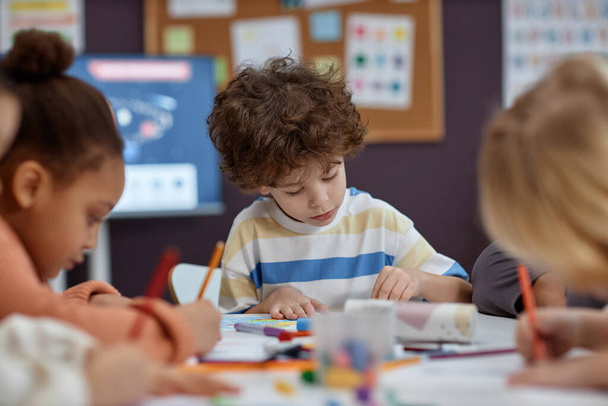 Portret van leuke jongen kleuren foto 's aan tafel met een groep kinderen in kunst en ambachten klasse in de kleuterschool, kopiëren ruimte - Foto, afbeelding