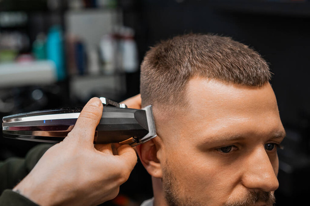 Barbeiro experiente usa aparador automático para cortar o cabelo do cliente no templo em barbearia closeup. Cabeleireiro faz penteado elegante por espelho de salão - Foto, Imagem