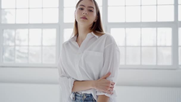 Gevşek beyaz bir gömlek ve kot pantolon giyen zarif genç bir kadın büyük pencereli, güneşli bir stüdyoda güvenle duruyor. Beyaz Gömlekli Genç Kadın Parlak Stüdyoda Gelişigüzel Poz Veriyor - Video, Çekim
