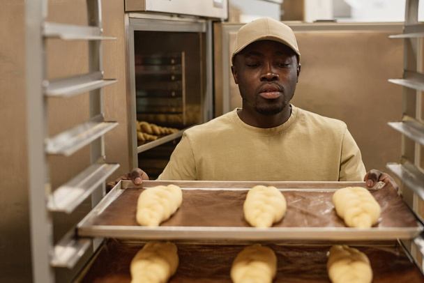 Visão frontal retrato do jovem negro como trabalhador na padaria artesanal segurando bandeja com pães frescos, espaço de cópia - Foto, Imagem