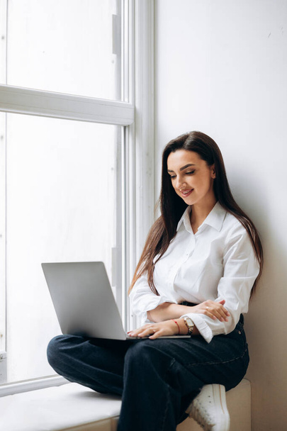 Bizneswoman siedzi na parapecie i używa laptopa. Kierownik finansowy pracujący w biurze. - Tak. Wysokiej jakości zdjęcie - Zdjęcie, obraz