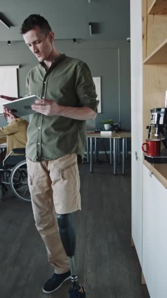 Κάθετη λήψη του ανθρώπου με το προσθετικό πόδι στέκεται στο γραφείο δίπλα στη μηχανή καφέ και χρησιμοποιώντας ψηφιακή ταμπλέτα κατά τη διάρκεια της εργάσιμης ημέρας - Πλάνα, βίντεο