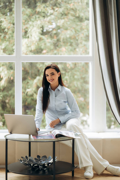 Gerente de design feminino atraente trabalhando no laptop na frente de uma grande janela. Lugar para trabalhar. Foto de alta qualidade - Foto, Imagem