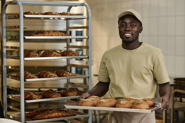 Porträt eines lächelnden Schwarzen, der Tablett mit frischem Brot in der Backküche in die Kamera lächelt, Kopierraum - Foto, Bild