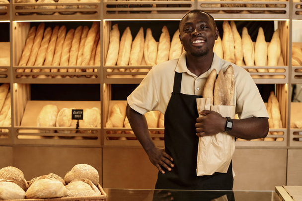 Retrato de cintura hacia arriba del joven negro parado detrás del mostrador en la panadería artesanal y sonriendo a la cámara, espacio para copiar - Foto, Imagen