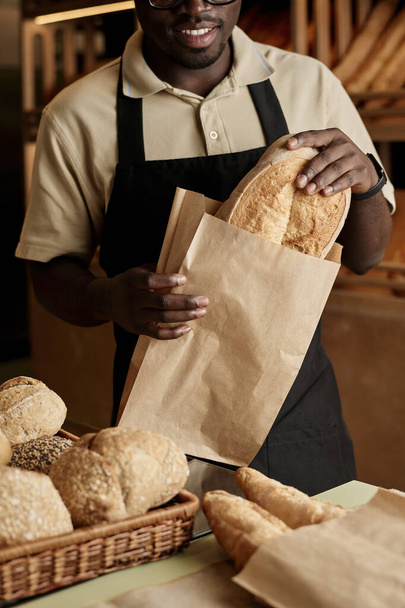 Κάθετη closeup του νέου άνδρα βάζοντας φρέσκο ψωμί σε χάρτινη σακούλα, ενώ εξυπηρετούν τον πελάτη σε βιοτεχνικό αρτοποιείο - Φωτογραφία, εικόνα