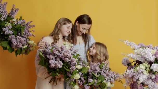 Nuori äiti halaa kahta lasta, näyttää ja hymyilee kameralle keltaisella taustalla luonnollisten kukkien kanssa studiossa. Onnellinen perhe. Tuki, liimaus tai hoito onnellinen äiti halaus, tytöt halaukset äiti. - Materiaali, video