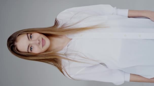 Un retrato de estudio de alta resolución de una mujer joven con el pelo largo y rubio y los ojos azules, con una camisa blanca. Retrato de una joven con una sonrisa suave en el estudio. Vídeo vertical. - Metraje, vídeo