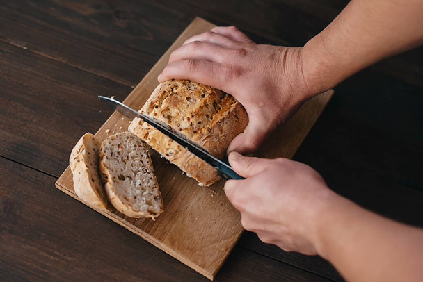 A person is slicing a bread loaf on a wooden cutting board. The staple food ingredient is a key component in various cuisine recipes - Photo, Image