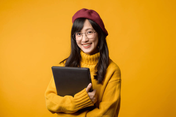 A laptopjával a kezében, fiatal ázsiai nő sárga pulóverben és piros sapkában magabiztosságot sugároz fényes sárga háttérrel szemben.. - Fotó, kép