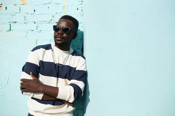 Portrait taille haute vibrant de jeune homme noir debout au mur bleu en ville portant des lunettes de soleil et tenue de style de rue, espace de copie - Photo, image