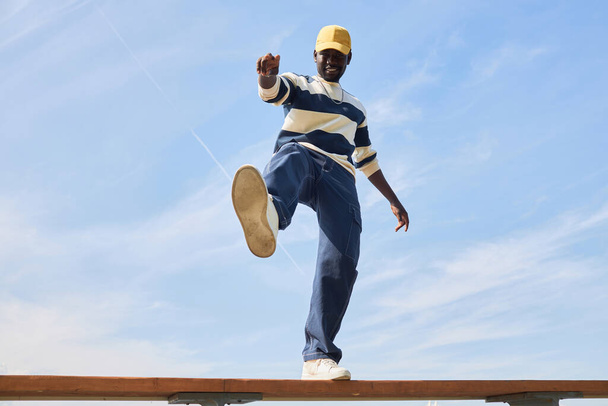 Prise de vue minimaliste de jeune homme noir portant une tenue tendance debout sur le toit contre le ciel bleu et donnant le coup d'envoi à la caméra. - Photo, image