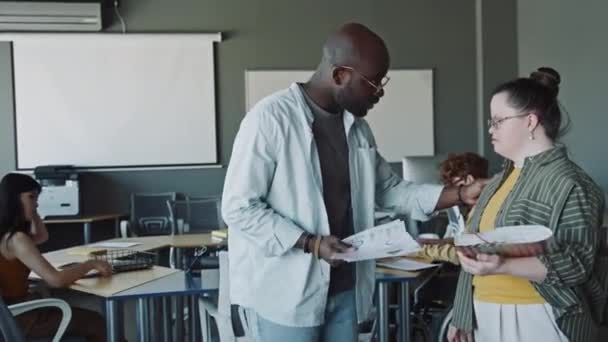 Афроамериканский менеджер приветствует молодую женщину-интерна с синдромом Дауна, показывает ей документы и объясняет бизнес-задачи во время рабочего дня в офисе - Кадры, видео