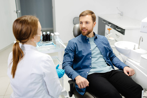 Blonde Zahnärztin in Zahnarztpraxis spricht mit männlichen Patienten und bereitet sich auf die Behandlung vor. Schöner bärtiger Mann im Zahnarztstuhl, der seinen Arzt lächelnd ansieht. - Foto, Bild