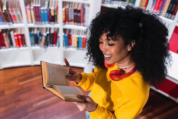 Высокий угол стильной счастливой молодой этнической студентки с длинными темными кудрявыми волосами в модном наряде и наушниками на шее, улыбающейся во время чтения книги в современной библиотеке - Фото, изображение