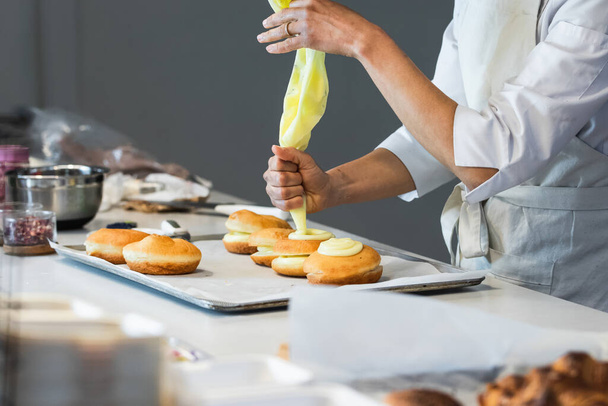 Вид сбоку на кукурузного анонимного пекаря, выдавливающего крем из кондитерской во время украшения сладких веганских берлинцев на кухне в пекарне - Фото, изображение