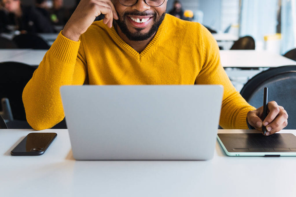 Imprenditore maschio afroamericano barbuto positivo irriconoscibile ritagliato che lavora su tablet grafico usando stilo e su netbook in ufficio - Foto, immagini