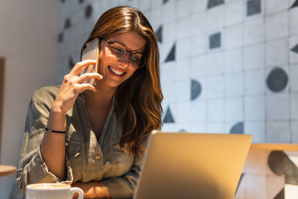 Joven mujer feliz en gafas hablando en el teléfono celular, mientras que en la mesa de café con netbook - Foto, Imagen