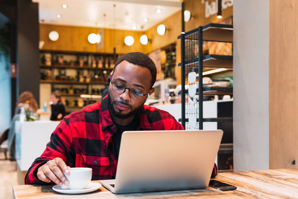 Позитивний афроамериканський бородатий чоловічий фрілансер переглядає нетбук з навушниками під час роботи віддалено в кафе і дивлячись на екран - Фото, зображення