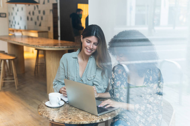 Através de parede de vidro vista de conteúdo multiétnico amigos do sexo feminino navegar na internet no netbook na mesa com xícara de café no café - Foto, Imagem
