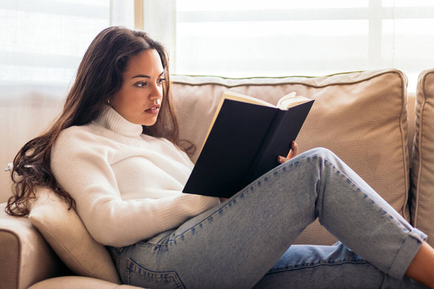 Evdeki kanepesinde oturan genç bir kadın, kendini bir kitaba kaptırmış. Okuma, rahatlama, iç huzur ve rahat bir ev. - Fotoğraf, Görsel