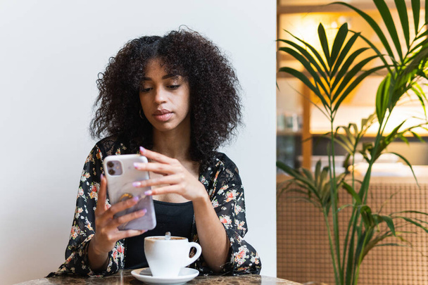カフェテリアテーブルで携帯電話でアフロヘアスタイルのブラウジングインターネットを持つ若い民族の女性,コーヒーのカップ - 写真・画像
