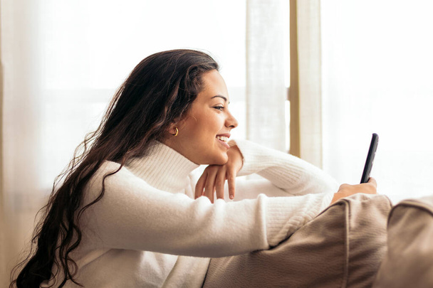 Mujer joven sentada en un cómodo sofá en su casa, usando su teléfono móvil con una expresión relajada en su cara. - Foto, imagen